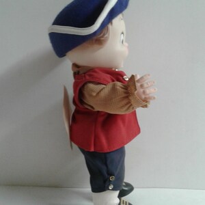 Pu&ograve; includere: Una bambola vintage vestita con un abito in stile coloniale. La bambola indossa un cappello blu con finiture bianche, un gilet rosso e pantaloni blu scuro. L'abito della bambola comprende una camicia color camoscio con maniche a balze, scarpe nere e calzini bianchi.