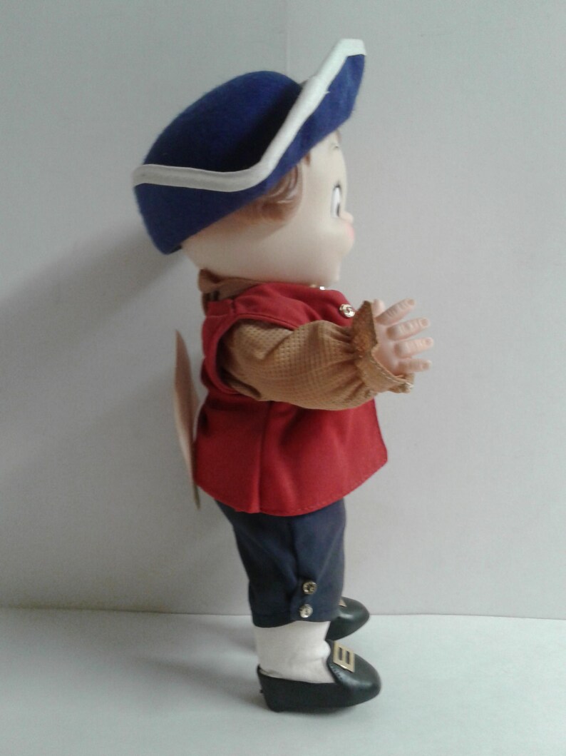 Pu&ograve; includere: Una bambola vintage vestita con un abito in stile coloniale. La bambola indossa un cappello blu con finiture bianche, un gilet rosso e pantaloni blu scuro. L'abito della bambola comprende una camicia color camoscio con maniche a balze, scarpe nere e calzini bianchi.