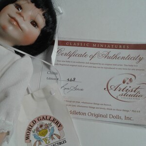 Pode incluir: Um certificado de autenticidade para uma boneca, com o texto "Classic Miniatures Certificate of Authenticity" no topo. O certificado &eacute; branco com texto preto e uma pequena imagem de uma pegada. A boneca &eacute; uma boneca de porcelana para beb&ecirc; com cabelo preto e um vestido branco. A boneca est&aacute; sentada em uma superf&iacute;cie branca. O certificado &eacute; assinado por "Seura Schick" e o n&uacute;mero da edi&ccedil;&atilde;o &eacute; 368.