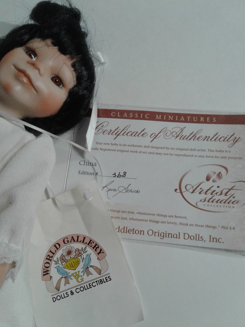 Pode incluir: Um certificado de autenticidade para uma boneca, com o texto "Classic Miniatures Certificate of Authenticity" no topo. O certificado &eacute; branco com texto preto e uma pequena imagem de uma pegada. A boneca &eacute; uma boneca de porcelana para beb&ecirc; com cabelo preto e um vestido branco. A boneca est&aacute; sentada em uma superf&iacute;cie branca. O certificado &eacute; assinado por "Seura Schick" e o n&uacute;mero da edi&ccedil;&atilde;o &eacute; 368.