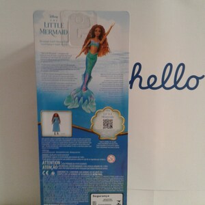 May include: The back of a doll's packaging featuring a mermaid doll with long red hair and a blue and green tail. The doll is wearing a blue and green top. The packaging is blue and features the words "Disney The Little Mermaid" and "Attention" in white text.