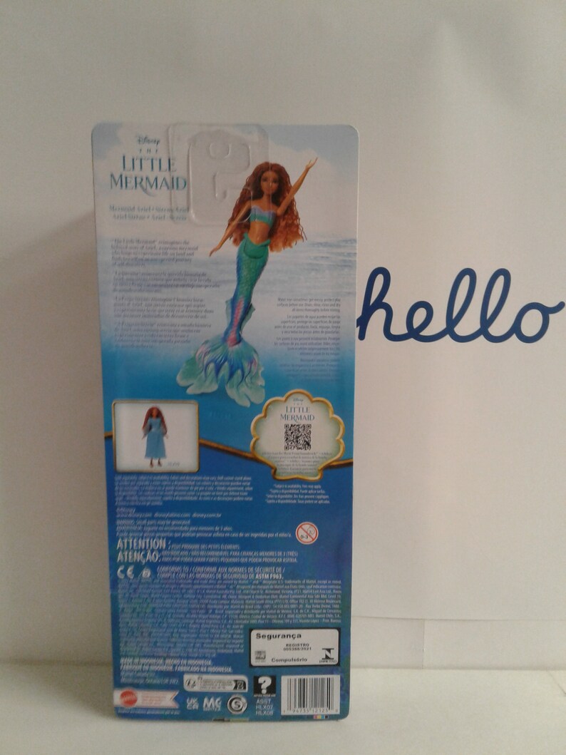 May include: The back of a doll's packaging featuring a mermaid doll with long red hair and a blue and green tail. The doll is wearing a blue and green top. The packaging is blue and features the words "Disney The Little Mermaid" and "Attention" in white text.