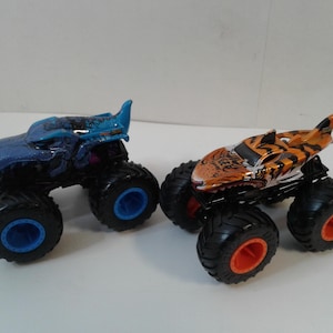 Może przedstawiać: Dwa zabawki monster truck. Jeden jest niebieski z niebieskimi kołami, drugi pomarańczowo-czarny z pomarańczowymi kołami. Oba mają duże, czarne opony i szczeg&oacute;łowe wzory.