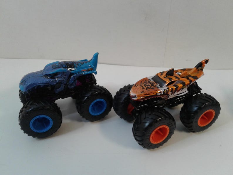 Może przedstawiać: Dwa zabawki monster truck. Jeden jest niebieski z niebieskimi kołami, drugi pomarańczowo-czarny z pomarańczowymi kołami. Oba mają duże, czarne opony i szczeg&oacute;łowe wzory.