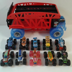 Może przedstawiać: Kolekcja zabawkowych monster truck&oacute;w w r&oacute;żnych kolorach i wzorach. Ciężar&oacute;wki mają duże koła i szczeg&oacute;łową grafikę. Duża czerwona ciężar&oacute;wka z niebieskimi kołami znajduje się na g&oacute;rze. Ciężar&oacute;wki są ułożone na białej powierzchni.