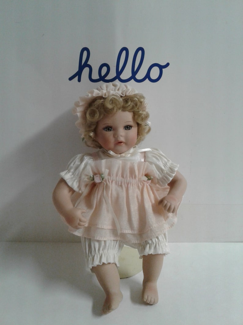 May include: A porcelain doll with blonde curly hair wearing a pink dress and white bloomers. The doll is sitting on a white surface with the word "hello" written in blue above it.