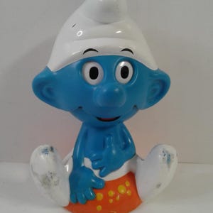 Pode incluir: Uma figura de um Smurf apresentando um personagem azul usando um chapéu branco e calças brancas, sobre uma base laranja decorada com pontos amarelos. O personagem tem olhos brancos grandes com pupilas pretas, nariz largo e um sorriso.