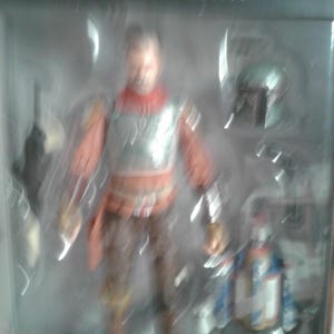 May include: A Star Wars The Mandalorian action figure in a plastic display case. The figure is wearing a brown and red outfit with a silver chest plate. The figure is holding a weapon in its right hand. The figure is standing on a black base. The text "Star Wars The Mandalorian" is printed on the base.