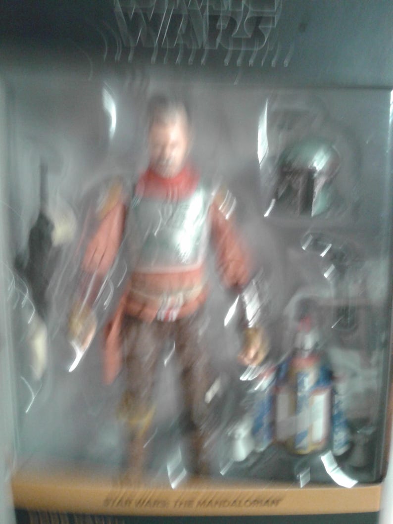 May include: A Star Wars The Mandalorian action figure in a plastic display case. The figure is wearing a brown and red outfit with a silver chest plate. The figure is holding a weapon in its right hand. The figure is standing on a black base. The text "Star Wars The Mandalorian" is printed on the base.