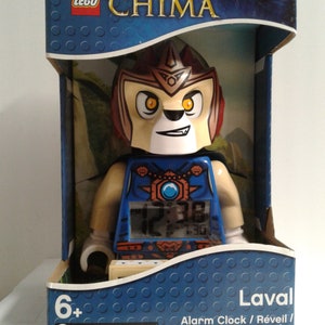 May include: A blue and brown LEGO Chima Laval alarm clock with a digital display. The clock features a LEGO minifigure of Laval, the lion warrior, with a blue and brown chest piece and a red and brown helmet. The clock is labeled "Laval" and "Alarm Clock/Réveil/Wecker/Despertador".