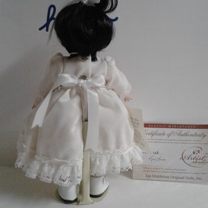 Pode incluir: Uma boneca de porcelana vestindo um vestido branco com detalhes em renda e sapatos brancos. A boneca tem cabelo preto preso em um rabo de cavalo e est&aacute; em p&eacute; em um suporte de metal. A boneca est&aacute; segurando um certificado de autenticidade que diz "Classic Miniatures Certificate of Authenticity" e &eacute; assinado por "Artist Studio Collection" e "Lee Middleton Original Dolls, Inc."