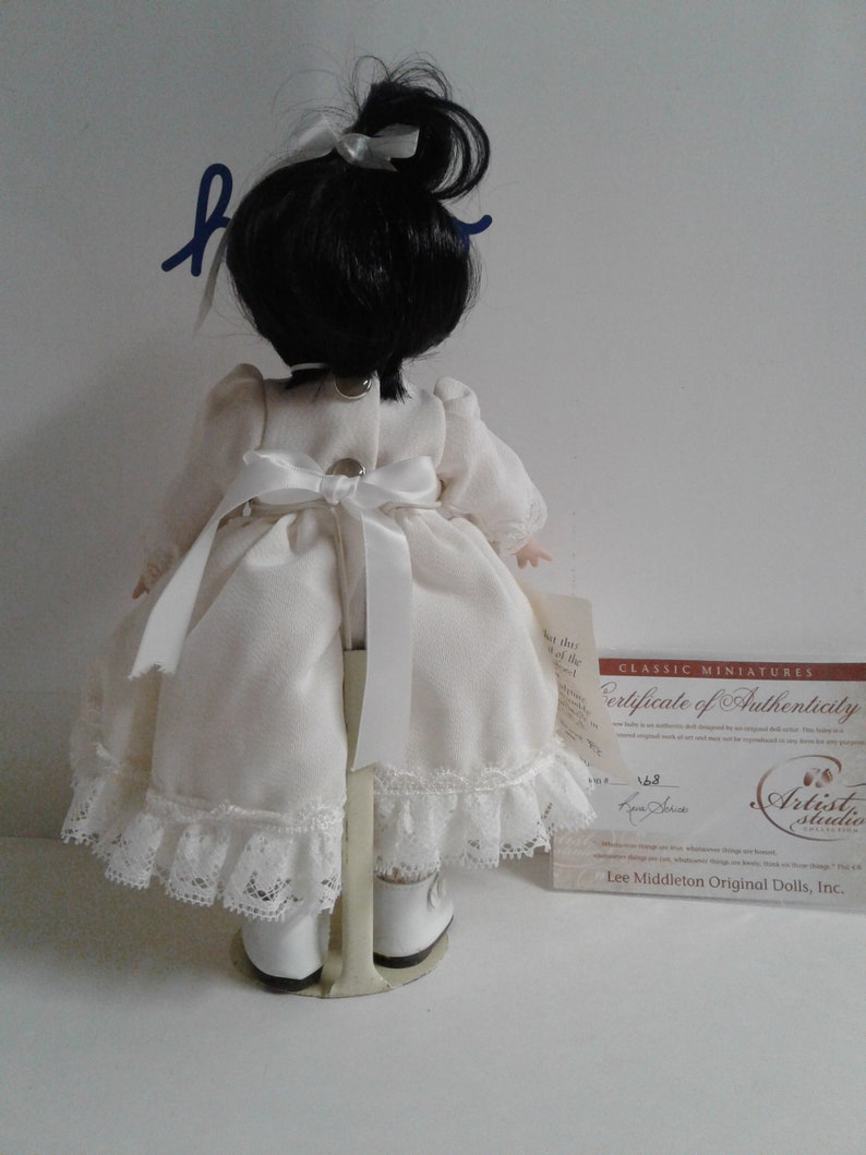 Pode incluir: Uma boneca de porcelana vestindo um vestido branco com detalhes em renda e sapatos brancos. A boneca tem cabelo preto preso em um rabo de cavalo e est&aacute; em p&eacute; em um suporte de metal. A boneca est&aacute; segurando um certificado de autenticidade que diz "Classic Miniatures Certificate of Authenticity" e &eacute; assinado por "Artist Studio Collection" e "Lee Middleton Original Dolls, Inc."