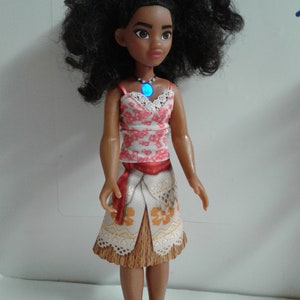 May include: A brown-skinned doll with long, curly black hair wearing a red and white floral top, a brown and white skirt with a red sash, and a blue necklace with a light blue gem.