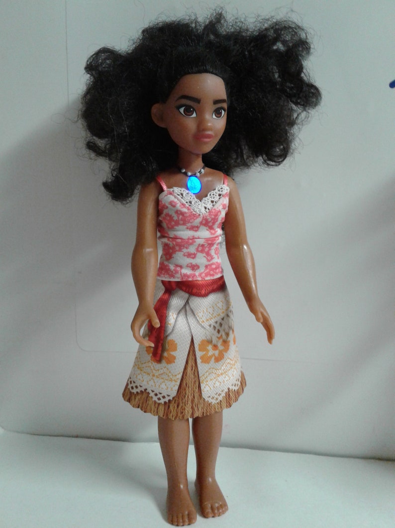 May include: A brown-skinned doll with long, curly black hair wearing a red and white floral top, a brown and white skirt with a red sash, and a blue necklace with a light blue gem.
