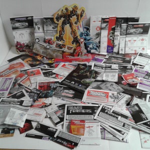 Könnte beinhalten: Ein großer Stapel von Transformers Spielzeug-Anleitungen in verschiedenen Farben, darunter Schwarz, Weiß, Rot und Blau. Die Anleitungen zeigen Bilder von Transformers-Charakteren und dem Transformers-Logo.