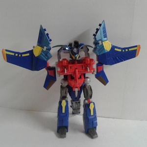 Peut inclure: Un robot jouet bleu, rouge et jaune avec de grandes structures en forme d'ailes s'étendant de son dos. Le robot présente un design détaillé avec de multiples articulations et un agencement complexe de pièces, suggérant une figurine transformable ou articulée.