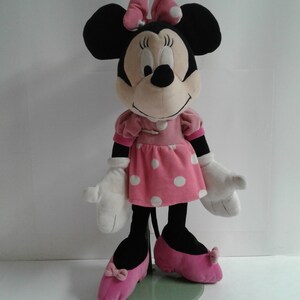 May include: A plush Minnie Mouse toy wearing a pink dress with white polka dots and pink shoes. She has a black nose and large black ears with a pink bow.