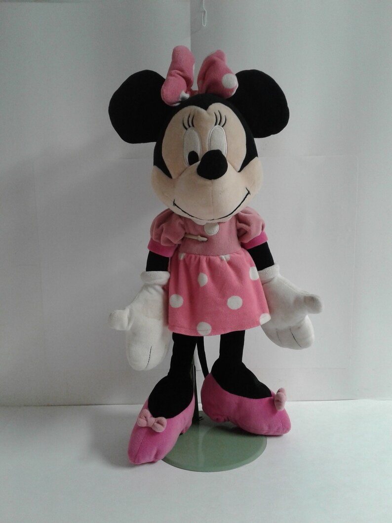 May include: A plush Minnie Mouse toy wearing a pink dress with white polka dots and pink shoes. She has a black nose and large black ears with a pink bow.