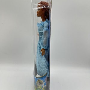 May include: A plastic-packaged doll with brown skin, long red hair, and a blue dress with white stripes. The doll is standing on a blue background with a fish and coral design.