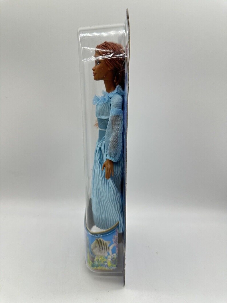 May include: A plastic-packaged doll with brown skin, long red hair, and a blue dress with white stripes. The doll is standing on a blue background with a fish and coral design.