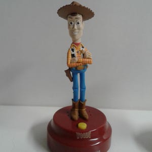 May include: A Woody figurine from the Toy Story franchise. The figure is wearing a brown cowboy hat, a yellow and orange plaid shirt, blue jeans, and brown boots. The figurine is standing on a red base with the name "Woody" and the words "ON" and "SENSOR" printed on it.