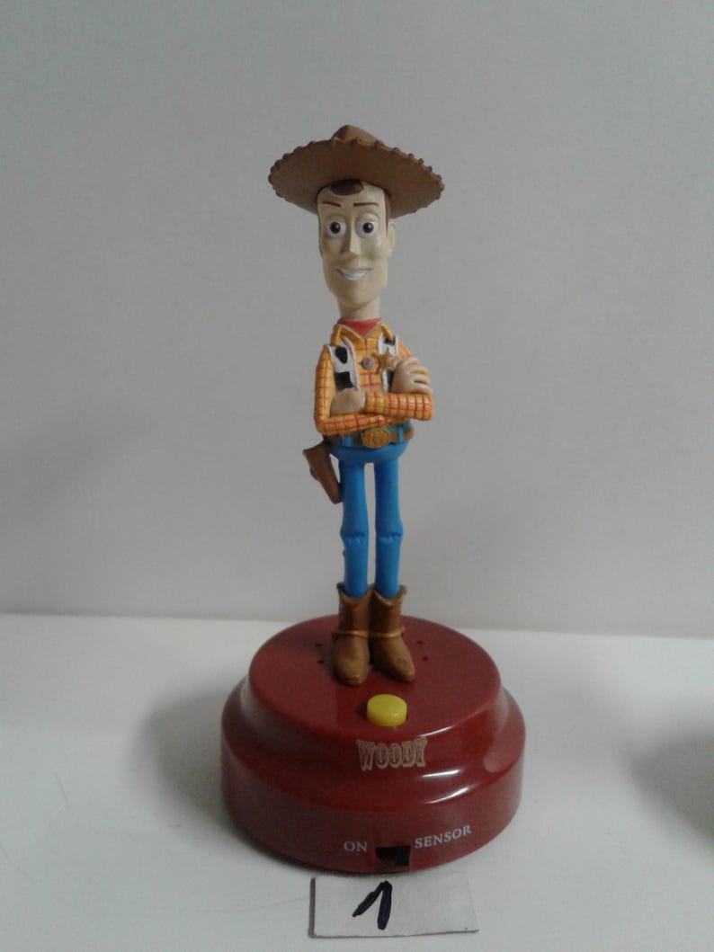 May include: A Woody figurine from the Toy Story franchise. The figure is wearing a brown cowboy hat, a yellow and orange plaid shirt, blue jeans, and brown boots. The figurine is standing on a red base with the name "Woody" and the words "ON" and "SENSOR" printed on it.