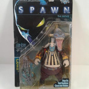 Peut inclure: Une figurine Spawn du film, encore dans son emballage d'origine. La figurine représente un personnage de clown avec une grosse tête et une bouche ouverte. L'emballage porte les mots "Spawn" et "The Movie".