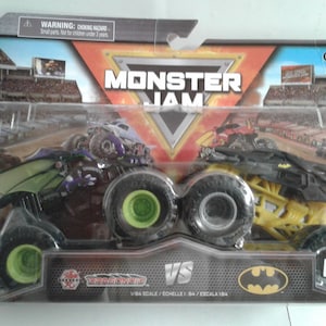 May include: Sealed Monster Jam toy set with two monster trucks. One truck is black and purple with a dragon design, the other is yellow and black with a Batman logo. The package features the text "Monster Jam" and "VS". Scale 1:64.