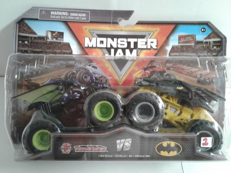 May include: Sealed Monster Jam toy set with two monster trucks. One truck is black and purple with a dragon design, the other is yellow and black with a Batman logo. The package features the text "Monster Jam" and "VS". Scale 1:64.