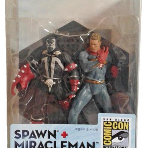 Może przedstawiać: Zestaw dwóch figurek akcji Spawn i Miracleman w oryginalnym opakowaniu. Figurka Spawn ma na sobie czarny kostium z białymi oznaczeniami i czerwone rękawice z kolcami. Figurka Miracleman ma na sobie niebieski kostium z czerwoną peleryną i czerwonymi rękawicami.