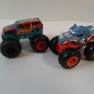 Może przedstawiać: Dwa zabawki monster truck. Jeden jest czerwony, zielony i ż&oacute;łty z numerem 68. Drugi jest niebieski i pomarańczowy z motywem rekina. Oba mają duże czarne opony z czerwonymi felgami.