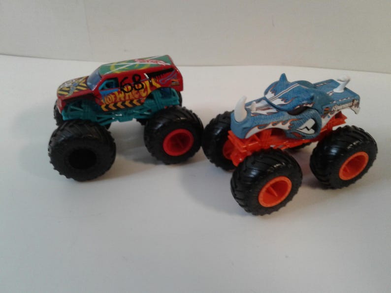 Może przedstawiać: Dwa zabawki monster truck. Jeden jest czerwony, zielony i ż&oacute;łty z numerem 68. Drugi jest niebieski i pomarańczowy z motywem rekina. Oba mają duże czarne opony z czerwonymi felgami.