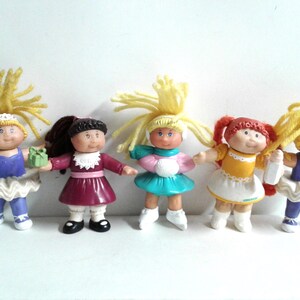 Könnte beinhalten: Fünf Vintage Cabbage Patch Kids Puppen. Jede Puppe hat eine einzigartige Frisur und ein Outfit, darunter Kleider in Lila, Rosa, Gelb und Türkis. Eine Puppe hält einen Stern, eine ein Geschenk und eine eine Flasche.