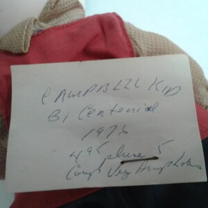 Pu&ograve; includere: Una bambola vintage con un gilet rosso e un maglione beige. Un biglietto scritto a mano &egrave; appuntato alla bambola, con il testo "Campbell Kid Bi Centennial 1976". I vestiti della bambola sono visibili.