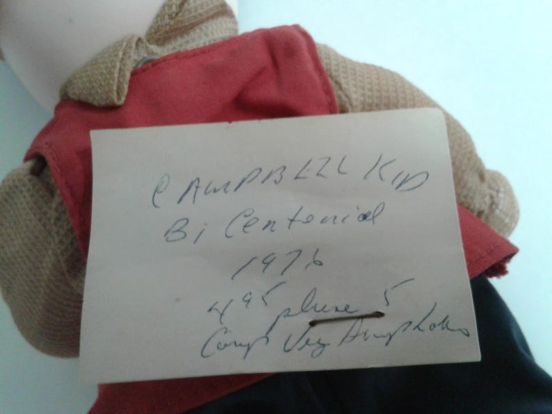Pu&ograve; includere: Una bambola vintage con un gilet rosso e un maglione beige. Un biglietto scritto a mano &egrave; appuntato alla bambola, con il testo "Campbell Kid Bi Centennial 1976". I vestiti della bambola sono visibili.