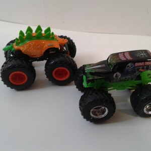 Może przedstawiać: Dwa zabawki monster truck. Jeden jest pomarańczowy z zielonymi kolcami i czerwonymi kołami. Drugi jest czarny i zielony ze słowami "Grave Digger" na g&oacute;rze i srebrnymi kołami.
