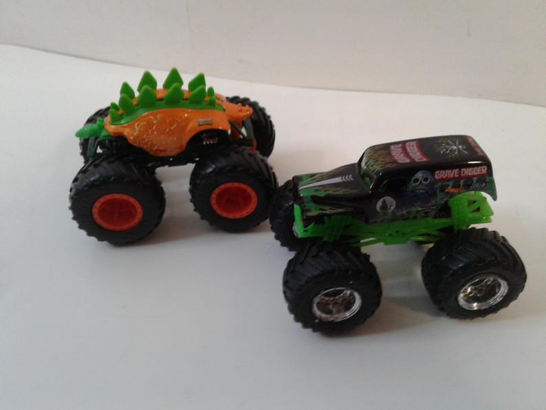 Może przedstawiać: Dwa zabawki monster truck. Jeden jest pomarańczowy z zielonymi kolcami i czerwonymi kołami. Drugi jest czarny i zielony ze słowami "Grave Digger" na g&oacute;rze i srebrnymi kołami.