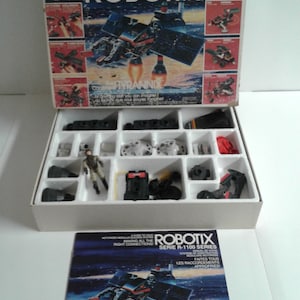 May include: A vintage ROBOTIX R-1100 Series toy construction set with a white box, a white foam tray with various parts, and a booklet with instructions. The box features a black and white illustration of a robot with the text "ROBOTIX R-1100 Series" and "The Motorized Modular Building System".
