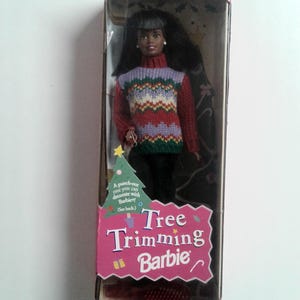 Może przedstawiać: Lalka Barbie Tree Trimming w oryginalnym opakowaniu. Lalka ma długie ciemne włosy i nosi dzianinowy sweter w czerwone, fioletowe, białe i zielone wzory oraz czarne spodnie. Pudełko zawiera grafikę choinki do wycięcia oraz napis Tree Trimming Barbie.