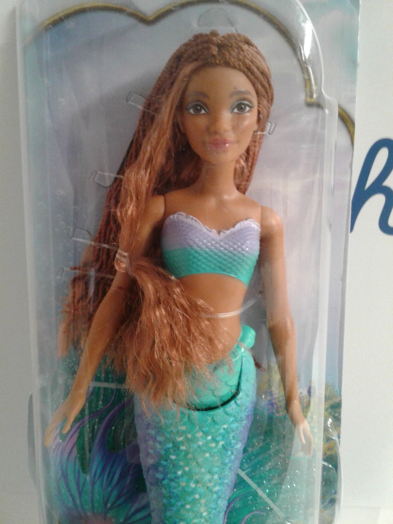 May include: A mermaid doll with long brown braided hair, wearing a teal and purple top and a teal mermaid tail with glitter accents.