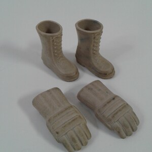 May include: A set of miniature tan-coloured combat boots and two miniature tan-coloured gloves. The boots have laces and a textured appearance. The gloves have detailed finger and wrist designs. The items are arranged on a white surface.