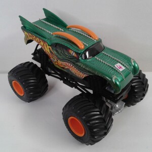 May include: Green and orange monster truck toy with large black tyres and orange rims. The truck features a dragon design on the hood and a number 4 on the side. The truck has a green body with orange accents.