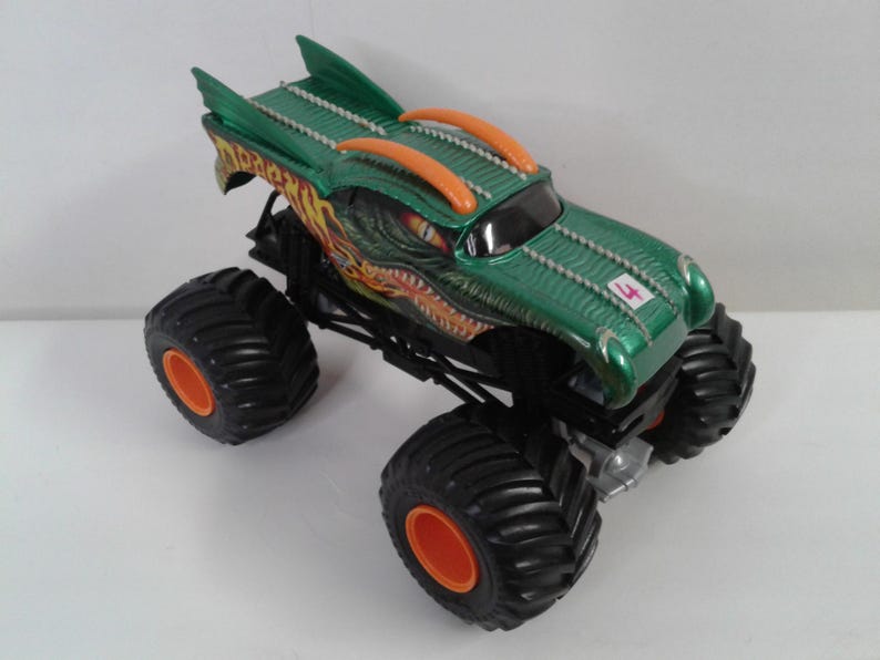 May include: Green and orange monster truck toy with large black tyres and orange rims. The truck features a dragon design on the hood and a number 4 on the side. The truck has a green body with orange accents.