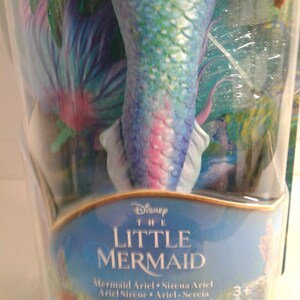 May include: A plastic package containing a mermaid doll with a blue, green, and pink tail. The doll is from the Disney movie The Little Mermaid. The package has the text "Disney The Little Mermaid" and "Mermaid Ariel Sirena Ariel Ariel Sirene Ariel - Sercia" on it. The package also has a Mattel logo and a 3+ age recommendation.