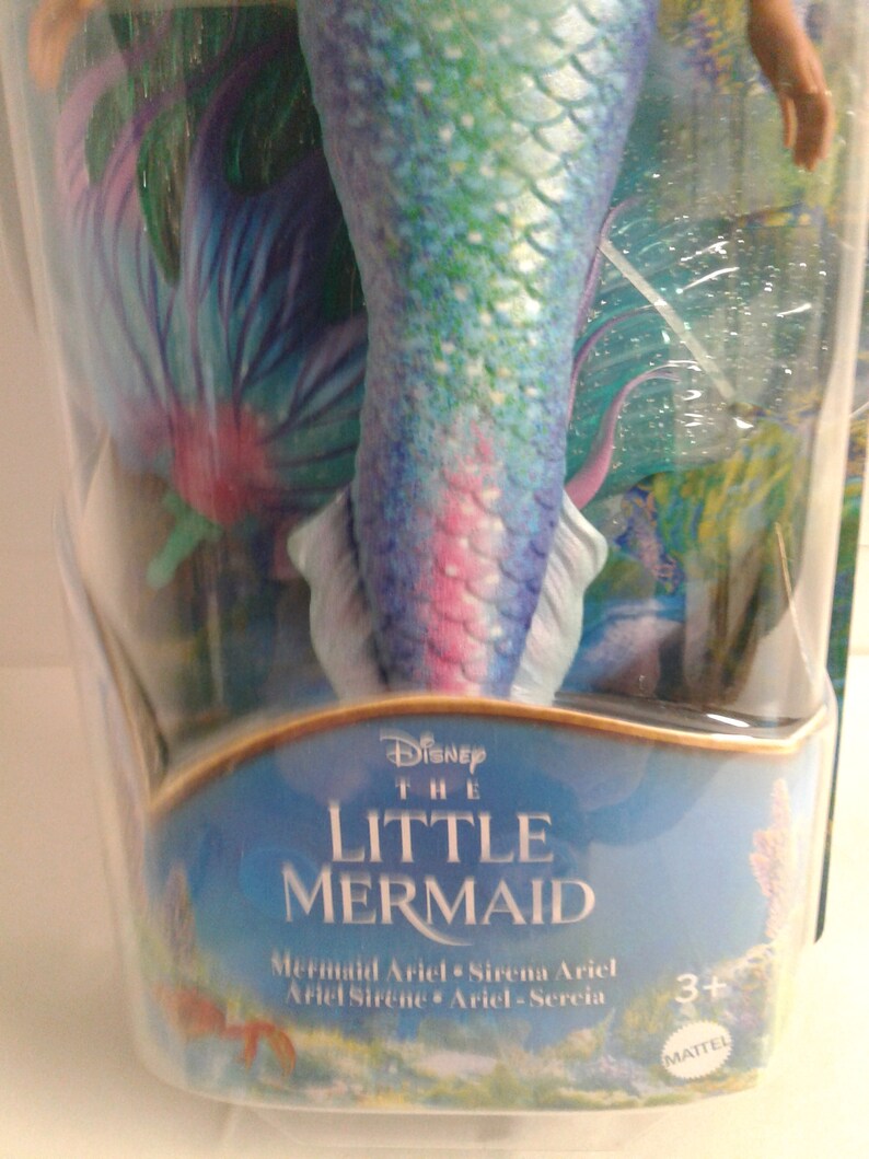 May include: A plastic package containing a mermaid doll with a blue, green, and pink tail. The doll is from the Disney movie The Little Mermaid. The package has the text "Disney The Little Mermaid" and "Mermaid Ariel Sirena Ariel Ariel Sirene Ariel - Sercia" on it. The package also has a Mattel logo and a 3+ age recommendation.