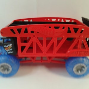 Może przedstawiać: Czerwono-czarna zabawka ciężar&oacute;wki Hot Wheels z niebieskimi kołami. Ciężar&oacute;wka ma czerwoną konstrukcję przypominającą klatkę i logo Hot Wheels. Koła są niebieskie ze srebrnymi felgami. Prz&oacute;d ciężar&oacute;wki ma niebiesko-czarny wz&oacute;r.