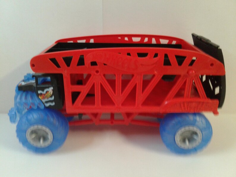 Może przedstawiać: Czerwono-czarna zabawka ciężar&oacute;wki Hot Wheels z niebieskimi kołami. Ciężar&oacute;wka ma czerwoną konstrukcję przypominającą klatkę i logo Hot Wheels. Koła są niebieskie ze srebrnymi felgami. Prz&oacute;d ciężar&oacute;wki ma niebiesko-czarny wz&oacute;r.