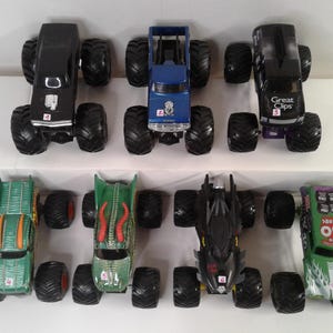 May include: A selection of toy monster trucks in various colours, including black, blue, green, and black. Each truck has large, black tyres and unique designs, some with logos and numbers.