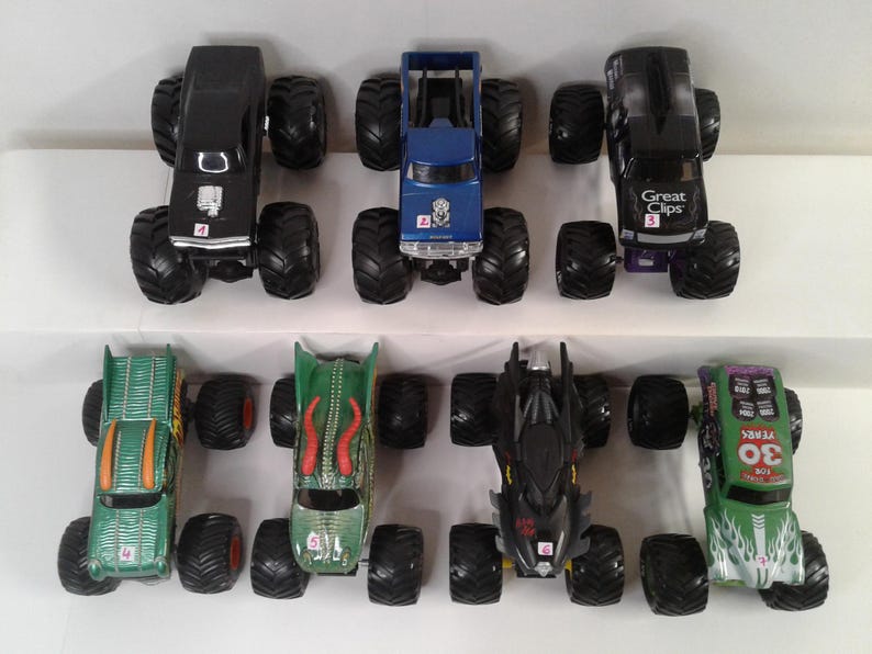 May include: A selection of toy monster trucks in various colours, including black, blue, green, and black. Each truck has large, black tyres and unique designs, some with logos and numbers.