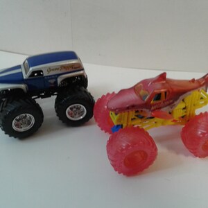 Może przedstawiać: Dwa zabawki monster truck. Jeden jest niebiesko-srebrny z czarnymi oponami. Drugi jest czerwono-ż&oacute;łty z czerwonymi kołami. Ciężar&oacute;wki zostały zaprojektowane z dużymi oponami i szczeg&oacute;łami.